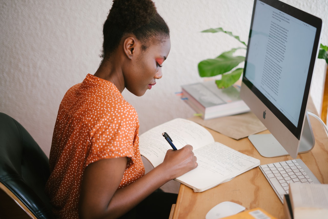 woman sat at a desk in front of a mac computer writing in a notebook