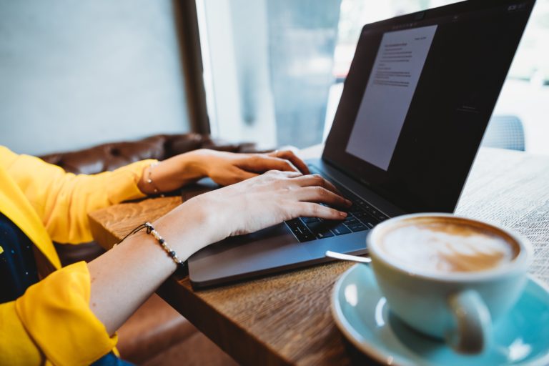 A person typing on their laptop in a cafe. They are wearing a yellow jacket and drinking a coffee.